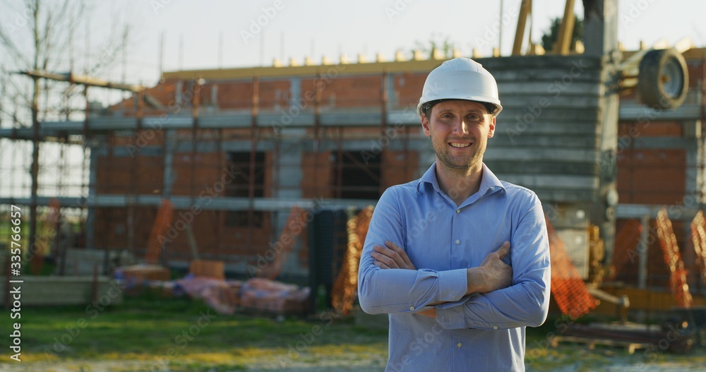 A young engineer is smiling in camera satisfied with his job on a background of construction building site. Concept: service, architecture, career people, construction A young engineer is smiling in camera satisfied with his job on a background of construction building site. Concept: service, architecture, career people, construction