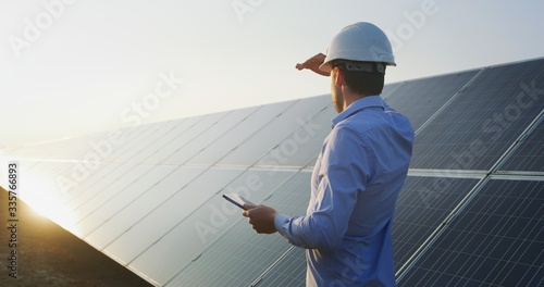 An young engineer is checking with tablet an operation of sun and cleanliness on field of photovoltaic solar panels on a sunset. Concept:renewable energy, technology,electricity,service, green,future 