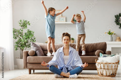 Happy mother with closed eyes meditating in lotus pose on floor trying to save inner harmony while excited children jumping on sofa and screaming in light spacious living room.