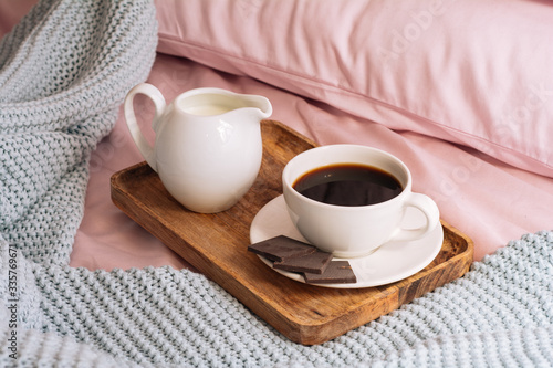 Breakfast in bed with freshly brewed and delicious coffee, a jug of cold milk and three slices of real bittersweet chocolate on a wooden tray against a backdrop of pink bedding and a blue plaid