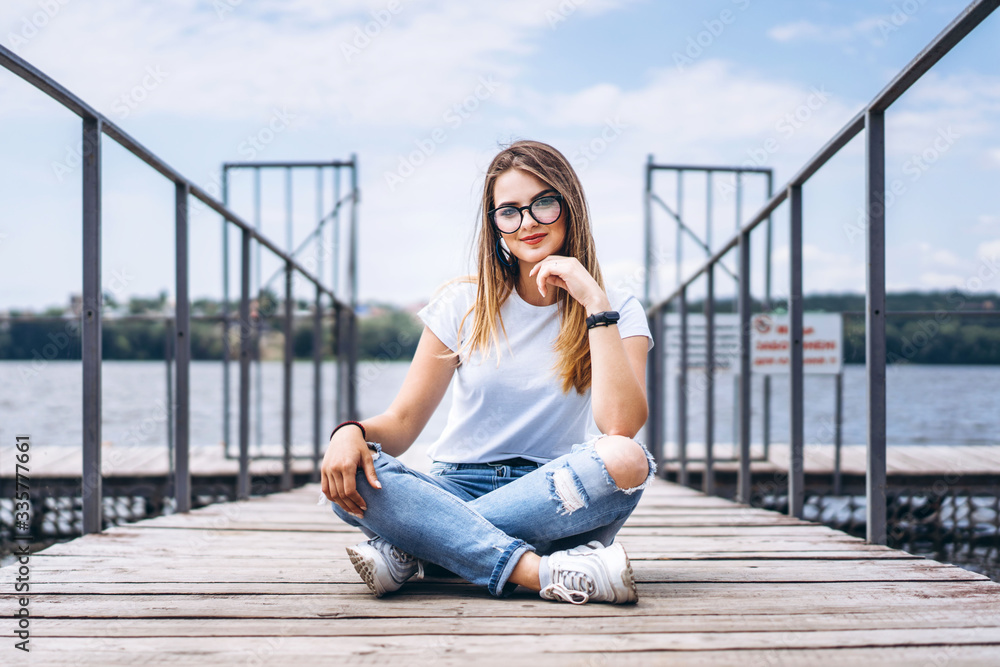 Young woman with long hair in stylish glasses posing on a wooden pier near the lake. Girl dressed in jeans and t-shirt smiling and looking at the camera