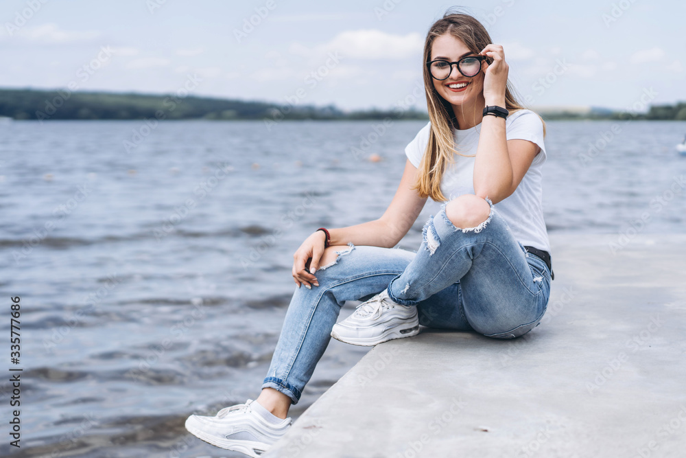 Young woman with long hair in stylish glasses posing on the concrete shore near the lake. Girl dressed in jeans and t-shirt smiling and looking at the camera Young woman with long hair in stylish glasses posing on the concrete shore near the lake. Girl dressed in jeans and t-shirt smiling and looking at the camera
