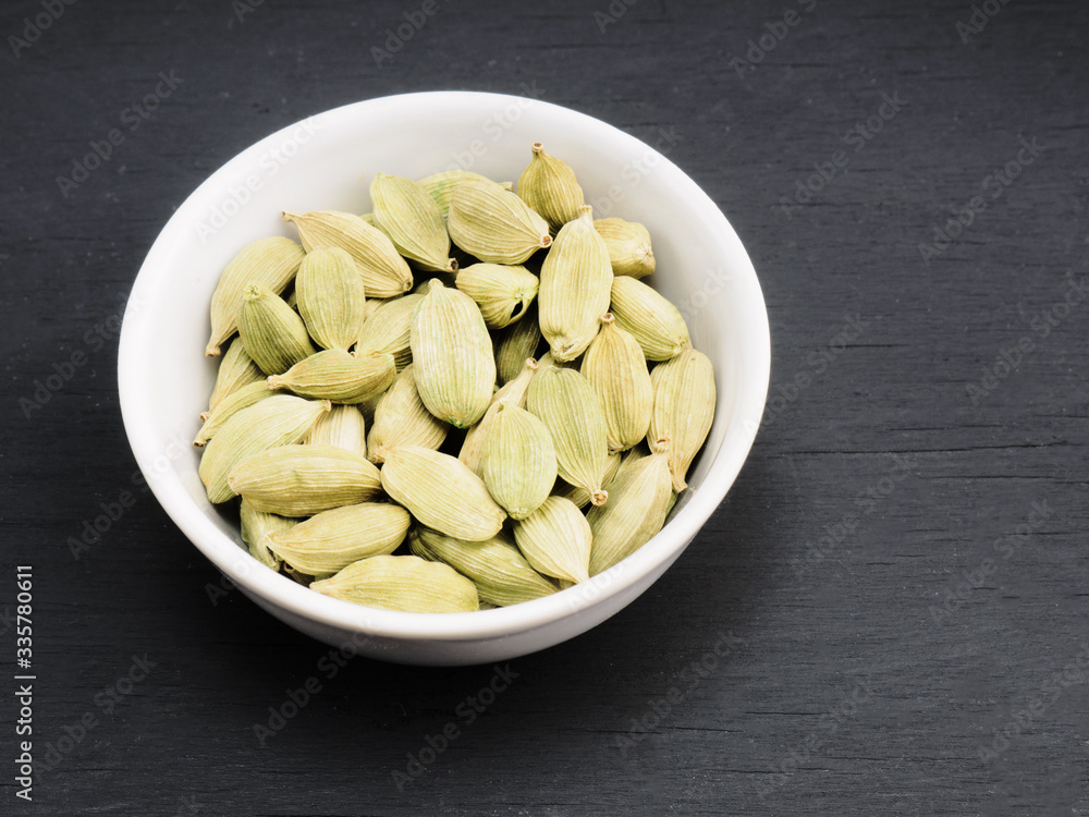 Spice green cardamom in white ceramic bowl on black wooden background. Healthy eating and organic food concept Spice green cardamom in white ceramic bowl on black wooden background. Healthy eating and organic food concept