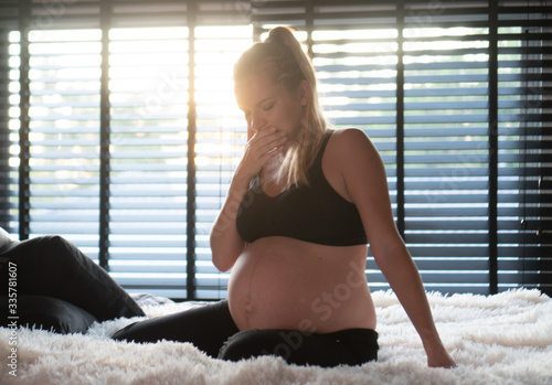 Pretty pregnant woman in black leggins and bra covering mouth with hand while sitting on her bed at home. Maternity, health care concept