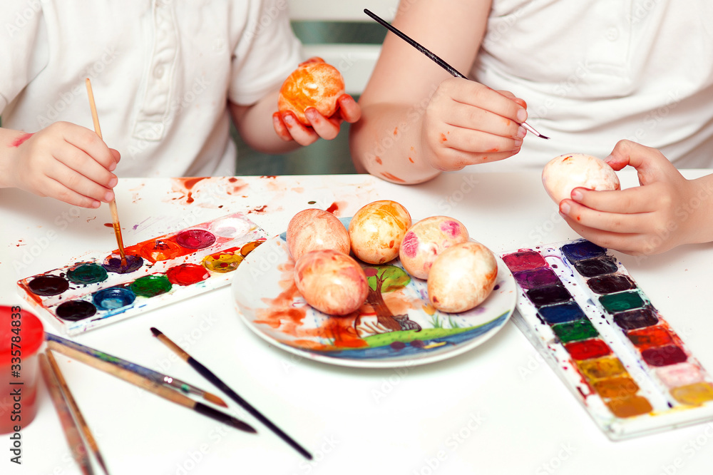 Bright watercolor paints lie on the table. Children's hands paint eggs. Easter