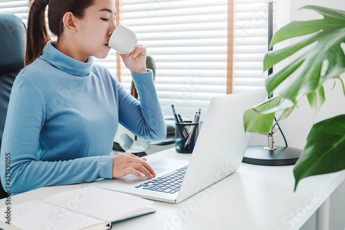 Asian woman working from home using computer and drinking coffee in her bedroom document finance and conference online meeting for new projects