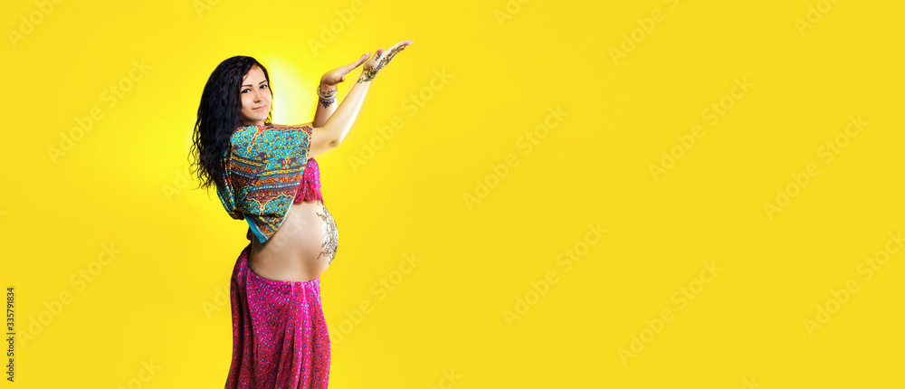 Pregnant happy woman in Indian sari with mehendi tattoo painted with henna on belly, fun fooling over yellow background, panoramic mock-up with space for text Pregnant happy woman in Indian sari with mehendi tattoo painted with henna on belly, fun fooling over yellow background, panoramic mock-up with space for text