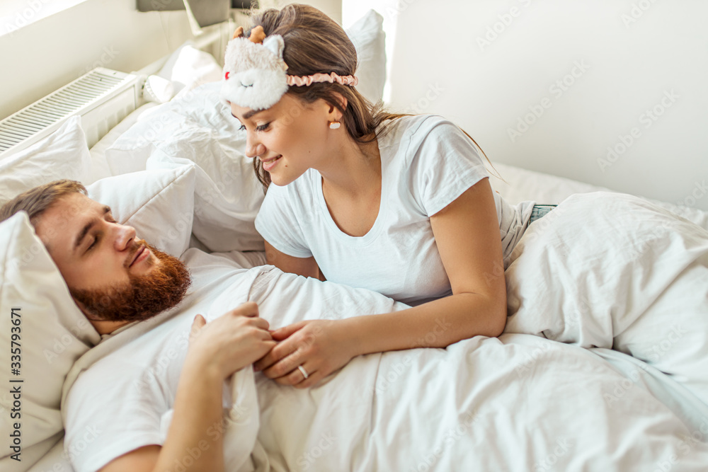 cute young caucasian woman look at sleeping man with love. married couple on bed in the morning. weekend, holiday, morning at home