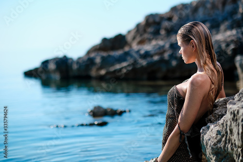 Young slim woman in bikini swimsuit looks at sea horizon. Happy woman relax and enjoying sun on the beach. Girl lying on big rocks sunbathing on the sea coast. Silhouette of slender woman on the beach © ZoomTeam