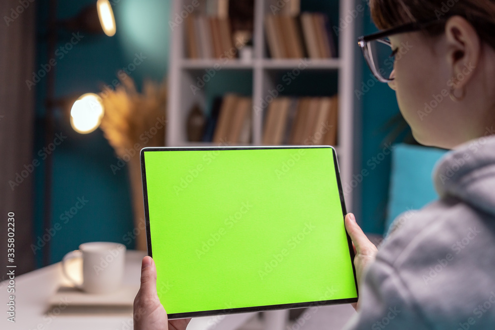 Back view of young female holding horizontal tablet with green screen sitting in room Back view of young female holding horizontal tablet with green screen sitting in room