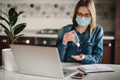 Coronavirus. Sanitizer gel. Business woman working, wearing protective mask in quarantine. Cleaning hands antibacterial gel to eliminate germs. Stay at home. Girl learns, using laptop. Freelance. photo