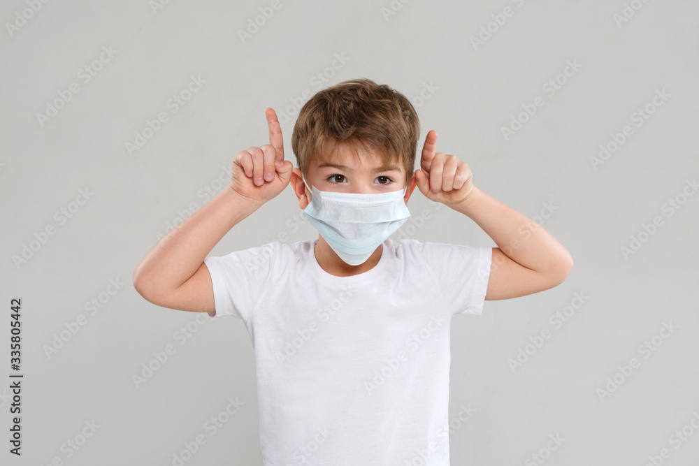 A little boy in a medical mask raised both hands to his head and depicts horns isolated on a gray background.