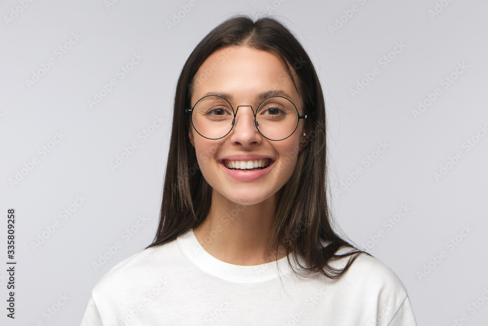 Close-up of young woman wearing big round glasses and white t-shirt smiling, laughing happily, looking at camera, isolated on gray background Close-up of young woman wearing big round glasses and white t-shirt smiling, laughing happily, looking at camera, isolated on gray background