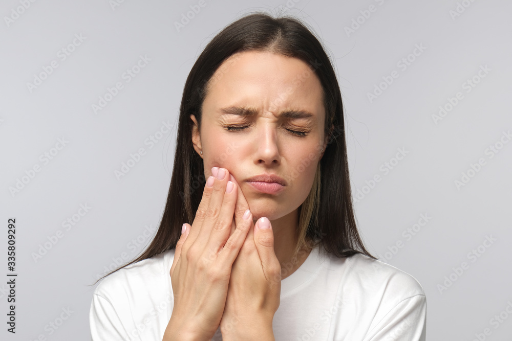 Young miserable woman experiencing severe toothache, pressing palm to cheek, closing eyes because of strong pain, isolated on gray background Young miserable woman experiencing severe toothache, pressing palm to cheek, closing eyes because of strong pain, isolated on gray background