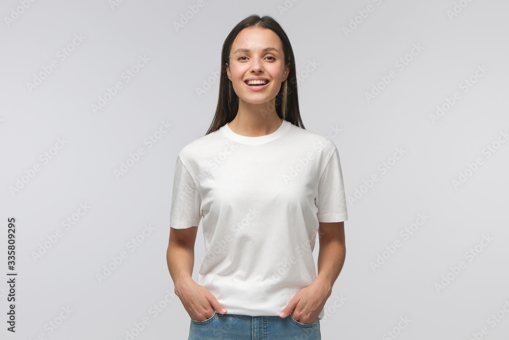 Laughing student girl wearing white T-shirt and blue jeans, standing with hands in pockets, looking straight at camera, isolated on gray background Laughing student girl wearing white T-shirt and blue jeans, standing with hands in pockets, looking straight at camera, isolated on gray background