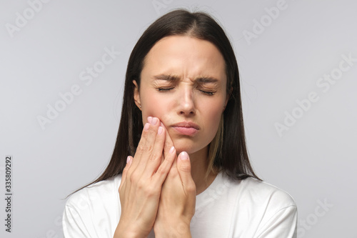 Young miserable woman experiencing severe toothache, pressing palm to cheek, closing eyes because of strong pain, isolated on gray background