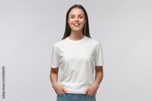 Laughing student girl wearing white T-shirt and blue jeans, standing with hands in pockets, looking straight at camera, isolated on gray background