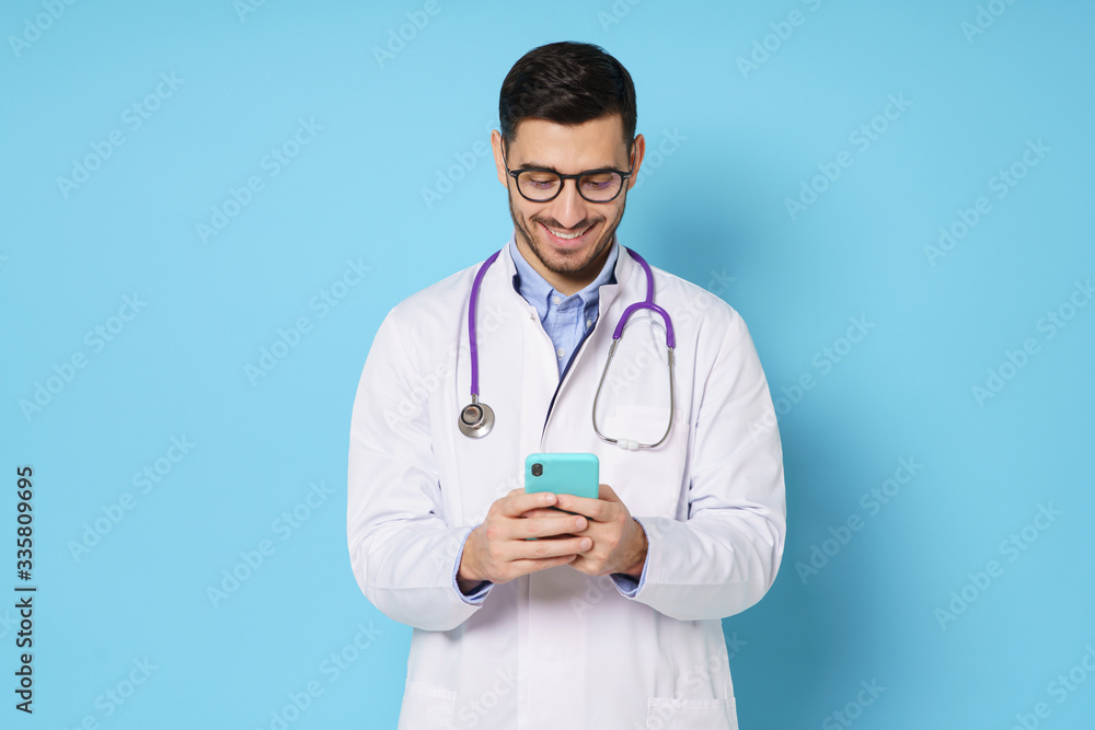 Young male doctor in white coat smiling while looking at screen of his phone, using medical app, standing isolated on blue background