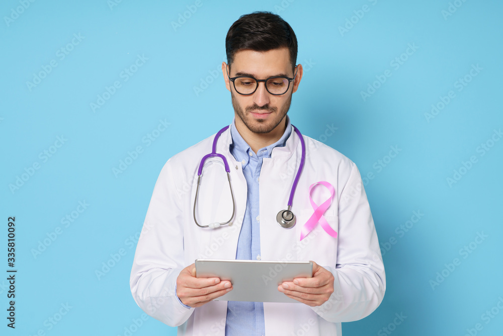 Young oncologist doctor with pink ribbon on his white coat looking at tablet, using app for medical check up, isolated on studio blue background. Breast cancer awareness month concept Young oncologist doctor with pink ribbon on his white coat looking at tablet, using app for medical check up, isolated on studio blue background. Breast cancer awareness month concept
