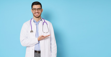 Horizontal banner of smiling young male doctor showing and presenting something with hand, isolated on blue background with copy space on right side
