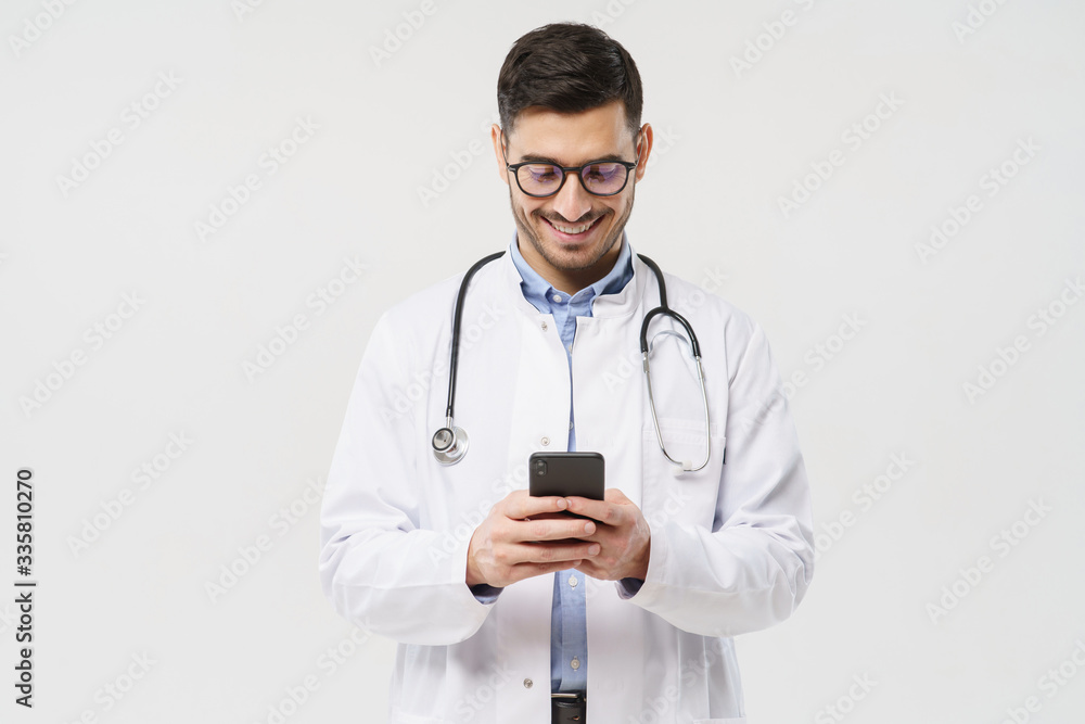 Young male doctor in white coat smiling while looking at screen of his phone, using medical app, standing isolated on gray background Young male doctor in white coat smiling while looking at screen of his phone, using medical app, standing isolated on gray background