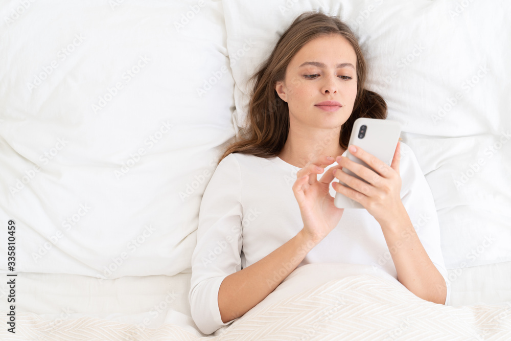 Young woman lying on bed pillows under blanket browsing social networks first time in morning, touching screen of smartphone with fingers Young woman lying on bed pillows under blanket browsing social networks first time in morning, touching screen of smartphone with fingers