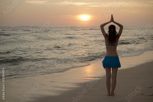 Yoga pose. Woman standing on the beach, practicing yoga. Young woman raising arms with namaste mudra during sunset golden hour. View from back. Melasti beach, Bali.