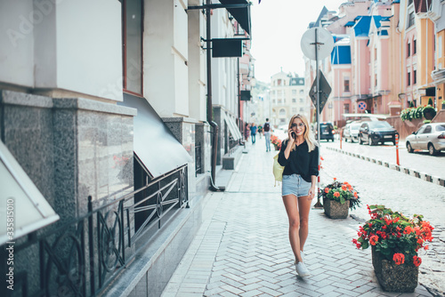 Full length portrait of cheerful female traveller exploring urban streets during summer vacations enjoying positive conversation, prosperous woman in trendy apparel connecting to roaming for calling © BullRun