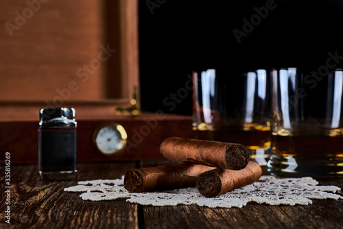 Still life with three cuban cigars, two glasses of whiskey or rum, lighter and wooden box with hygrometer. Old wooden table top and black background.