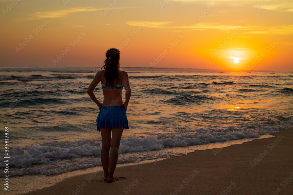 Young Caucasian woman walking along the beach. Sunset golden hour. Travel concept. View from back. Copy space. Melasti beach, Bali, Indonesia.