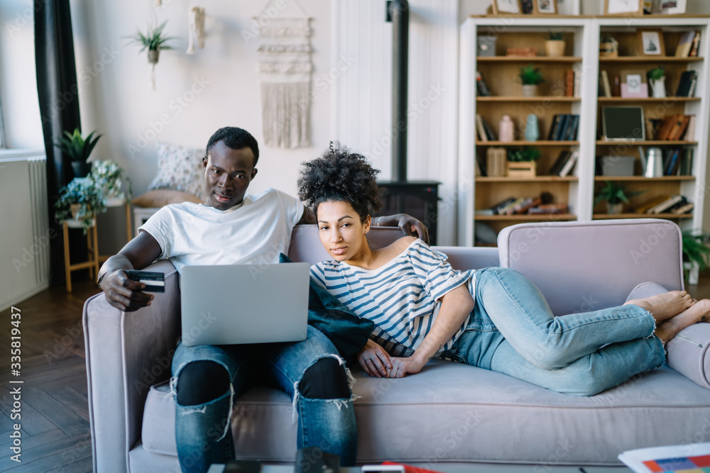 Confident multiracial couple placing in sofa with credit card and laptop Confident multiracial couple placing in sofa with credit card and laptop