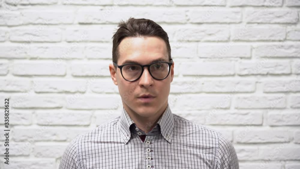 Handsome young Caucasian brunet man businessman in black glasses in a checkered gray shirt looks away, nods, agrees with someone against white brick wall background close up. Handsome young Caucasian brunet man businessman in black glasses in a checkered gray shirt looks away, nods, agrees with someone against white brick wall background close up.
