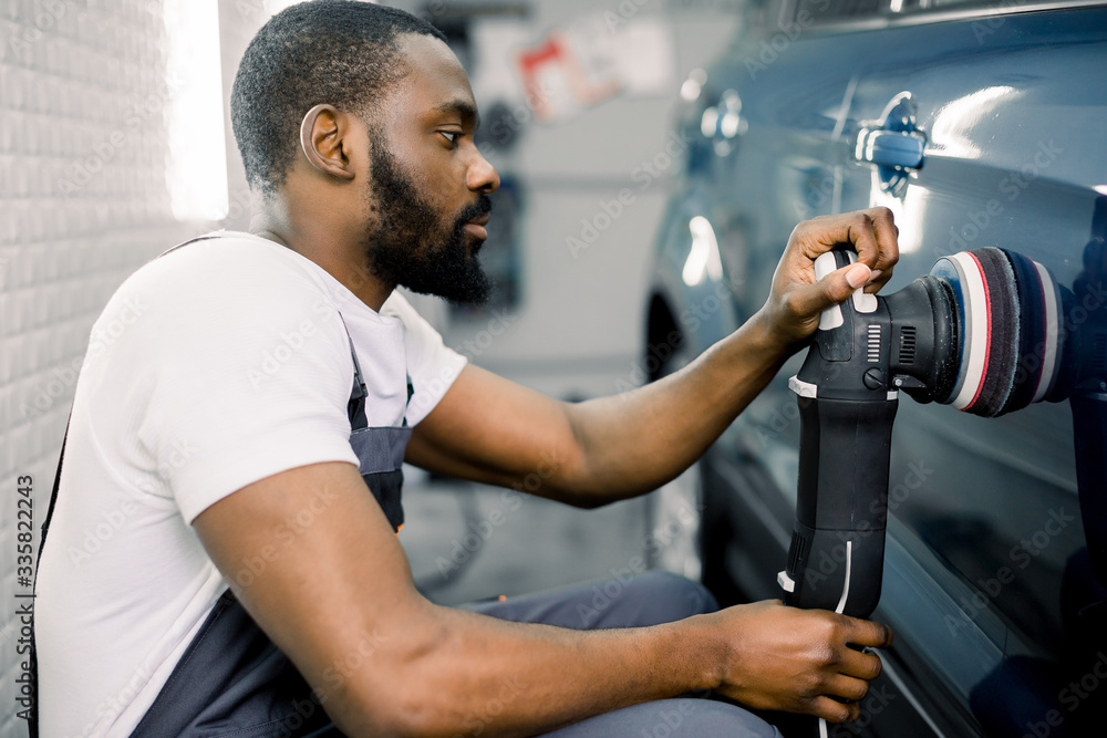 Auto detailing service, polishing of the car. Side view of young African American man worker n t-shirt and overalls, polishing blue car door with orbital polisher Auto detailing service, polishing of the car. Side view of young African American man worker n t-shirt and overalls, polishing blue car door with orbital polisher