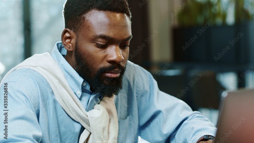 Obraz premium Portrait of african american handsome male using laptop for work in modern office programmer serious looks at monitor screen freelance successful worker businessman close up