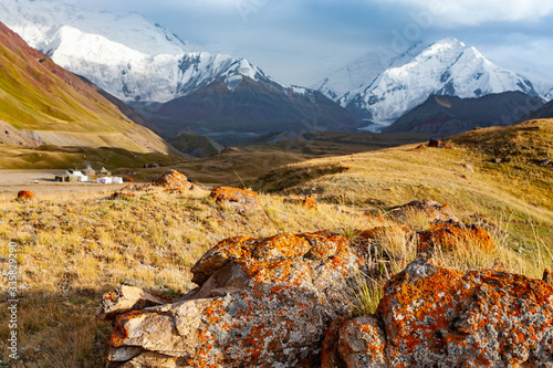 Stone with orange moss and lichen on foreground. Snowy mountains. Beautiful landscape of Kyrgyz nature. Tent camp. photo