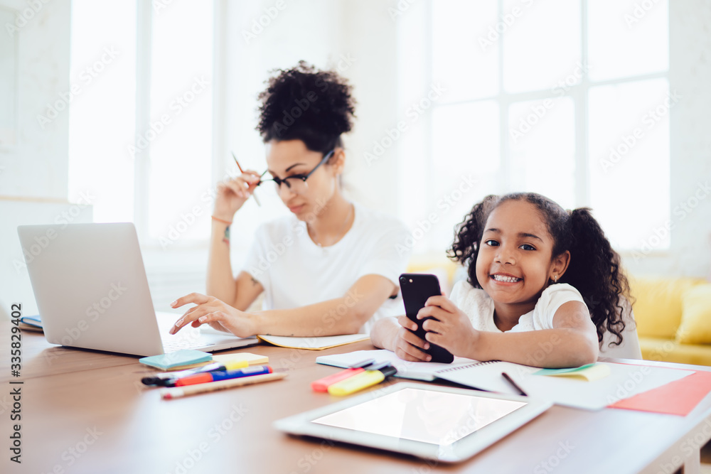 Blurred mother using laptop and daughter playing on smartphone while looking at camera Blurred mother using laptop and daughter playing on smartphone while looking at camera
