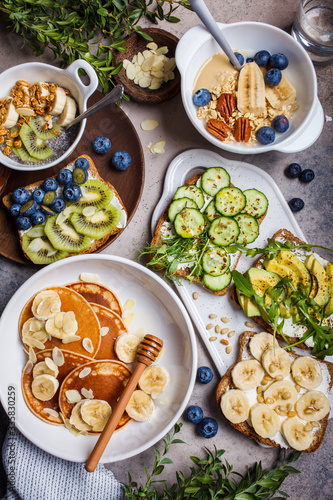 Healthy vegetarian breakfast, flat lay. Oatmeal with fruits, chia pudding, pancakes with banana and honey and toasts with fruits, vegetables and cream cheese.