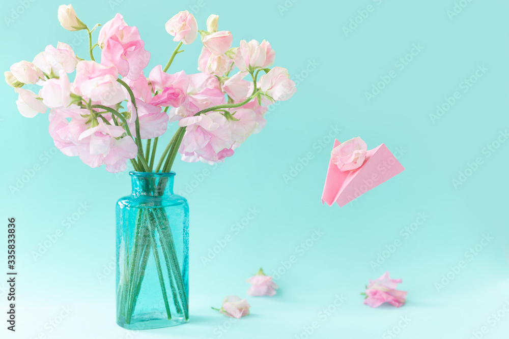 Obraz premium Bouquet of pink sweet pea Lathyrus in beautiful transparent vase on blue background.Paper airplane with message. Spring concept. International Women's Day, March 8 and Valentine's Day,14 of February