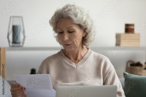 Focused serious old mature woman sitting on sofa, looking through paper documents, reading email letter, checking bank loan payment notification, managing medical insurance alone in living room.