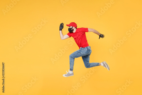 Fun jumping delivery man in red cap t-shirt uniform sterile face mask gloves isolated on yellow background studio Guy employee courier Service quarantine pandemic coronavirus virus 2019-ncov concept. © ViDi Studio