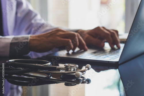 Male doctor in white coat typing on laptop computer keyboard with medical stethoscope on the desk in medical room at clinic or hospital. Online medical technology, emr, telehealth or meditech concept. photo