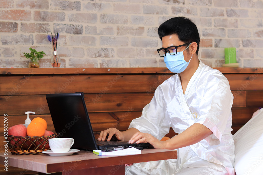 Asian Man working from home wearing protective mask in bedroom. wearing protective mask at home. Having sanitizer gel to get his hands cleaned. Working from home concept and drinking coffee Asian Man working from home wearing protective mask in bedroom. wearing protective mask at home. Having sanitizer gel to get his hands cleaned. Working from home concept and drinking coffee