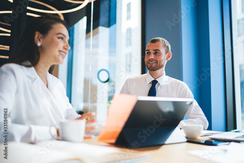 Cheerful male and female colleagues enjoying friendly brainstorming during working time in office, happy man and woman sitting at desk and smiling while discussing business ideas for project