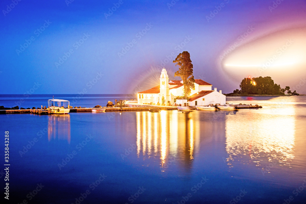 Beautiful charming optimistic view of Vlacherna Monastery on the Kanoni peninsula in Kerkyra, Corfu, Greece at night with a streak of light from airplane lamps in the sky.