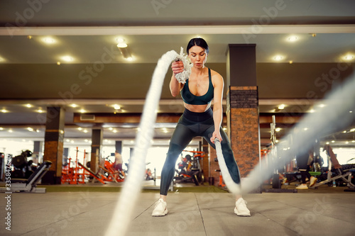 Caucasian fit woman dressed in sportsoutfit doing workout with battle ropes at gym. © zamuruev