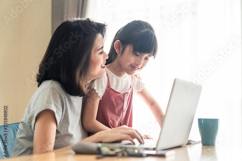 Asian young mother work from home. Woman sit and use laptop for meeting with colleague while daughter stand by her. Mom happy to do job while taking care family. Covid-19, Social distancing concept.