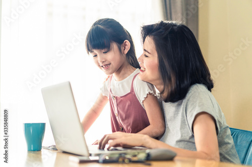Asian young mother work from home. Woman sit and use laptop for meeting with colleague while daughter stand by her. Mom happy to do job while taking care family. Covid-19, Social distancing concept.