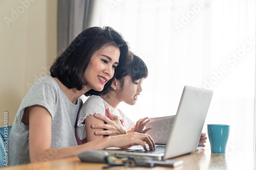 Asian young mother work from home. Woman use laptop for meeting with colleague while daughter sit ,be hug from behind. Mom happy to do job while taking care family. Covid-19, Social distancing concept