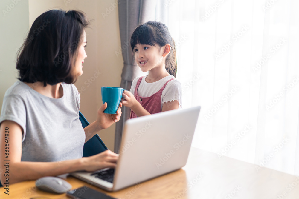 Asian young office mother work from home during coranavirus outbreak. Cute daughter give a cup of coffee to mom and kiss for a break and rest. Love care in family. Covid-19, Social distance concept. Asian young office mother work from home during coranavirus outbreak. Cute daughter give a cup of coffee to mom and kiss for a break and rest. Love care in family. Covid-19, Social distance concept.