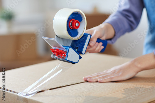 Close up of unrecognizable young woman using tape dispenser for packing boxes while moving out or relocating, copy space photo